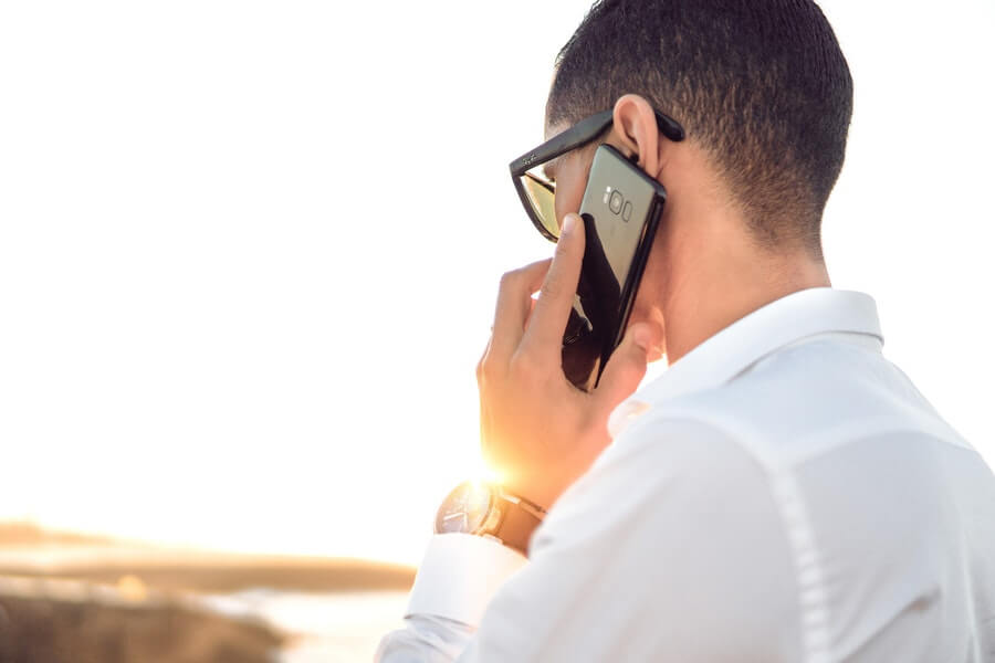 Man who looks like a professional or businessman wearing a watch and a white dress shirt talking on an Android phone on the beach looking at the ocean.