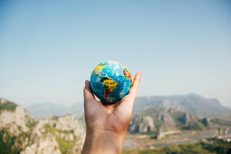 Hand (with wrist) holding small globe ball in the air against scenic background.