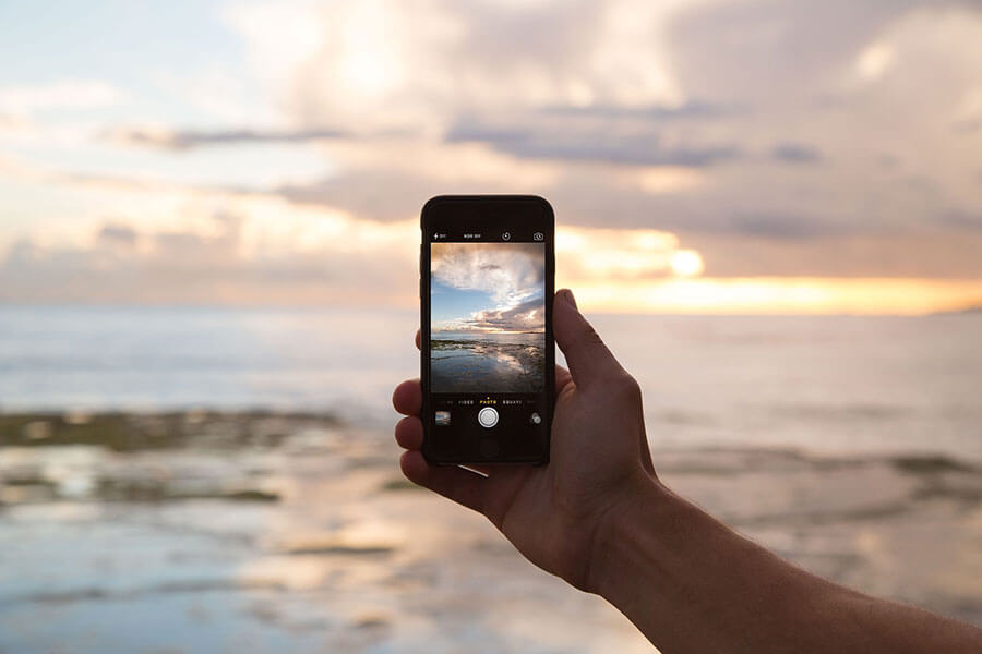 Somebody's hand holding a phone taking a picture of the ocean during a beautiful sunrise/sunset on the beach.