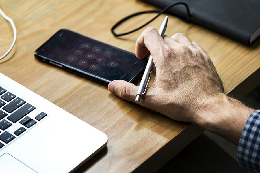 Man's hand with a pen in it grabbing iPhone with a laptop and notebook on a desk.