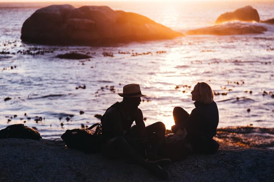 Man and woman (probably a couple) sitting on a beach at sunset/sunrise, looking at each other against the beautiful ocean in the background.