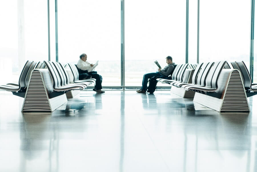 Two men sitting across one another at the airport, reading newspapers.