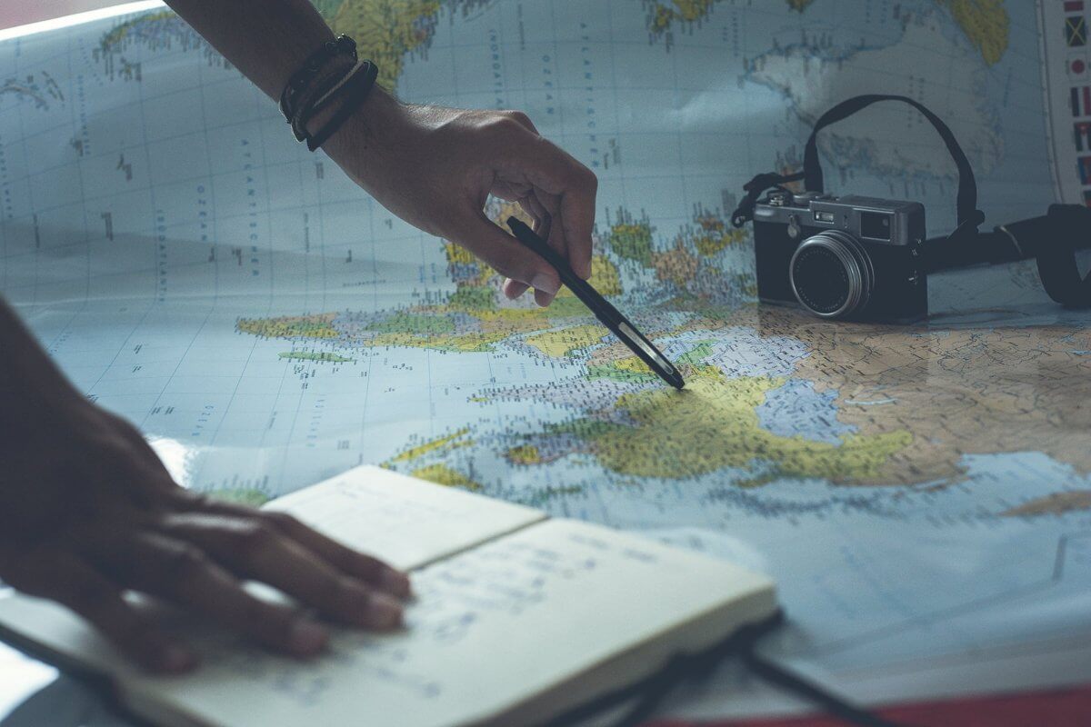 Camera and notebook on a map, with a hand holding a pencil, planning a trip.