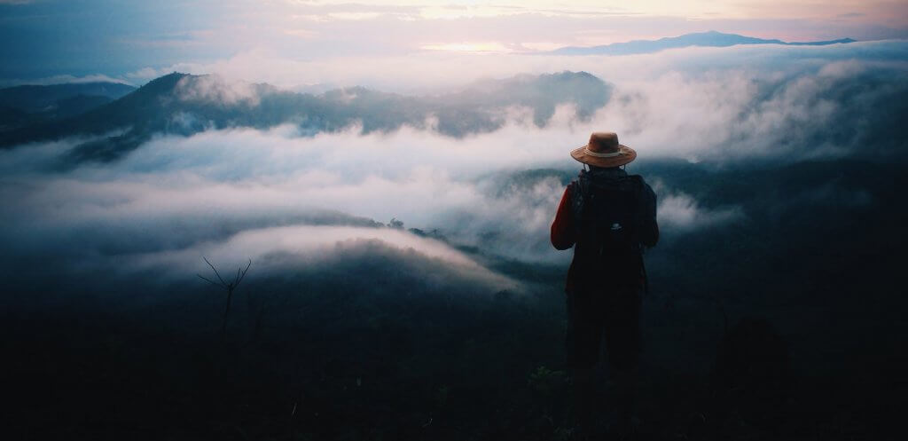 Single person standing alone on a mountain above the clouds with a backpack and hat.