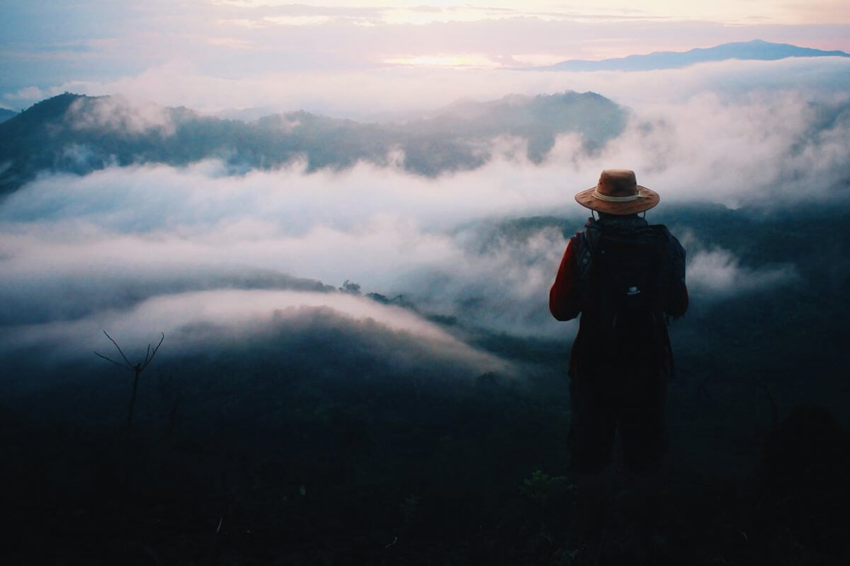 Single person standing alone on a mountain above the clouds with a backpack and hat.